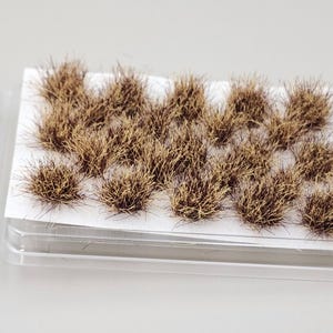 May include: Miniature model terrain grass tufts in a clear plastic container. The grass is a mix of brown and tan fibers. A small, gray figurine of a person playing a stringed instrument is also included.
