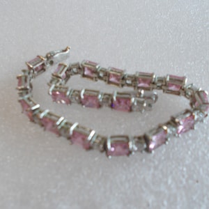 May include: A silver-toned link bracelet featuring a series of square-cut pink stones alternating with small clear accent stones. The bracelet is designed with a secure metal clasp mechanism and is arranged in a circular shape on a textured surface.