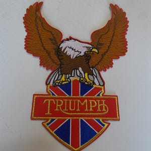May include: Embroidered patch featuring a brown eagle with outstretched wings above a red banner with the word "TRIUMPH" in gold lettering. The banner is above a shield with a Union Jack design.
