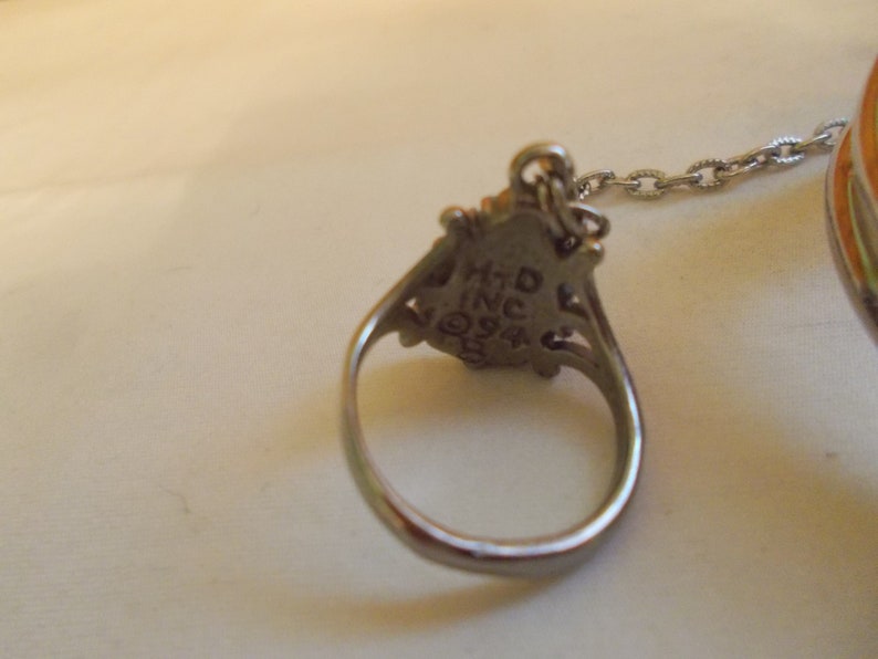 May include: A silver ring with a decorative design and a chain attached. The ring has the inscription "HTD INC 924" on the back.