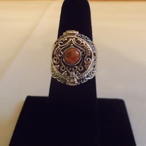 May include: A silver ring with an ornate design and a small, round, orange stone in the center. The ring is a hinged box that opens to reveal a small compartment.