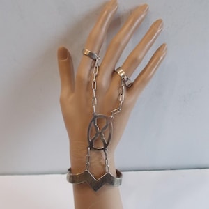May include: A silver hand chain with two rings connected by a chain to an oval-shaped centerpiece. The bracelet has a zig-zag design. The jewelry is displayed on a mannequin hand.