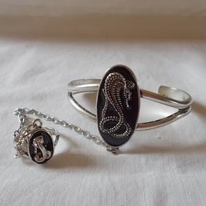 Könnte beinhalten: Ein silbernes Armband und ein Ringset mit einem schwarzen Schlangenmotiv aus Emaille. Das Armband ist offen und der Ring ist mit einer Kette am Armband befestigt.
