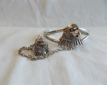 May include: Silver-toned jewelry set featuring an eagle design. The set includes a cuff bracelet and a pendant with a chain. The eagle heads have detailed feathering and a realistic appearance. The jewelry is displayed on a white surface.