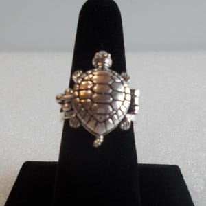May include: A silver-toned ring featuring a detailed turtle motif on the face. The turtle shell has a segmented, rounded design with visible head, tail, and flipper details. The metal has a polished finish with darkened recessed areas.