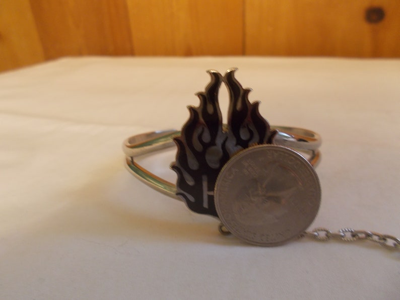 May include: Silver bracelet with a black and white flame design. The bracelet has a chain attached to it. A US quarter is included for size reference.