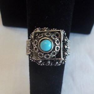 May include: A silver ring with a square filigree design and a turquoise stone in the center.