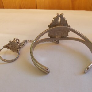 May include: A silver bracelet and ring set with a decorative design. The bracelet is open and has a chain connecting it to the ring.