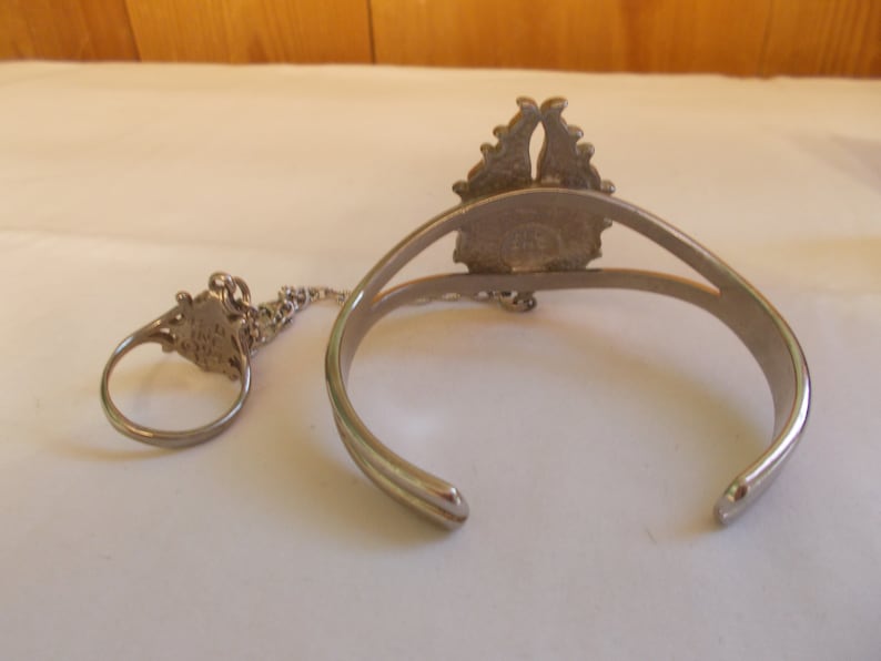 May include: A silver bracelet and ring set with a decorative design. The bracelet is open and has a chain connecting it to the ring.