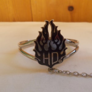 May include: A silver bracelet with a black and silver flame design and the letters "HD" in the center. The bracelet has a chain attached to it.