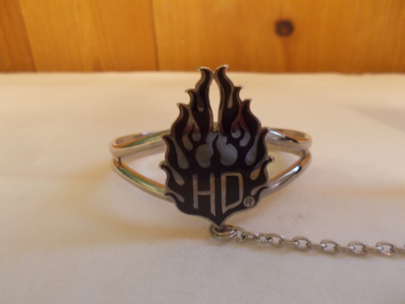 May include: A silver bracelet with a black and silver flame design and the letters "HD" in the center. The bracelet has a chain attached to it.