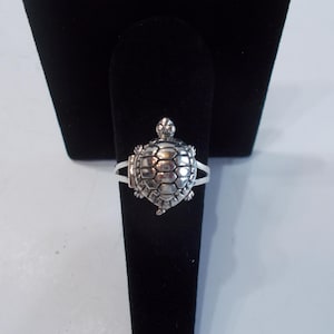 May include: A silver-colored ring featuring a detailed turtle design. The turtle's shell is textured, and the ring has a split shank. The ring is displayed on a black velvet jewelry stand, showcasing its intricate details.