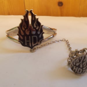 May include: A silver bracelet and ring set with a black and silver flame design and the letters "HD" in the center. The bracelet has a chain connecting it to the ring.