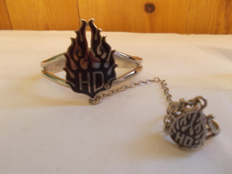 May include: A silver bracelet and ring set with a black and silver flame design and the letters "HD" in the center. The bracelet has a chain connecting it to the ring.