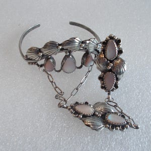 May include: A silver-toned cuff bracelet with a connected ring. The bracelet features shell-shaped accents and teardrop-shaped pink stones. A chain connects the bracelet to a ring with similar shell and stone designs.
