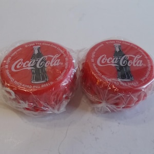 Może przedstawiać: Dwa czerwone plastikowe jo-jo w kształcie zakrętki od butelki Coca-Coli. Każde jo-jo ma białe logo Coca-Coli i czarną sylwetkę butelki. Jo-jo są zapakowane w przezroczyste plastikowe opakowanie.