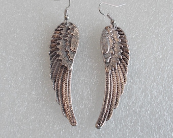 May include: A pair of silver-colored wing earrings. Each earring features a detailed wing design with layered feathers. The earrings have a hook closure for pierced ears. The wings have a metallic finish and are displayed against a light background.