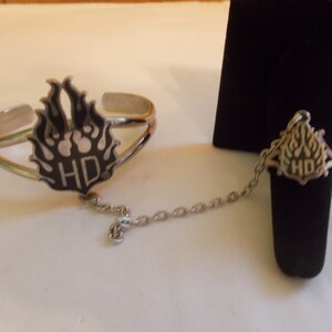 May include: A silver bracelet and ring set with a black and silver Harley Davidson logo. The bracelet and ring are connected by a silver chain.