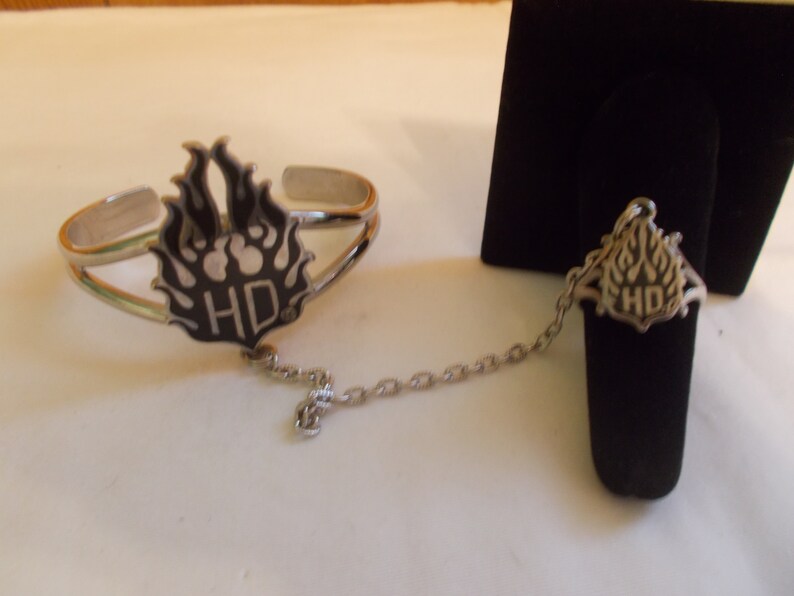 May include: A silver bracelet and ring set with a black and silver Harley Davidson logo. The bracelet and ring are connected by a silver chain.
