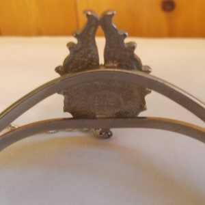 May include: Silver tone bracelet with a decorative design featuring a crown and a chain. The back of the bracelet has a circular stamp with the text "LMC" inside.