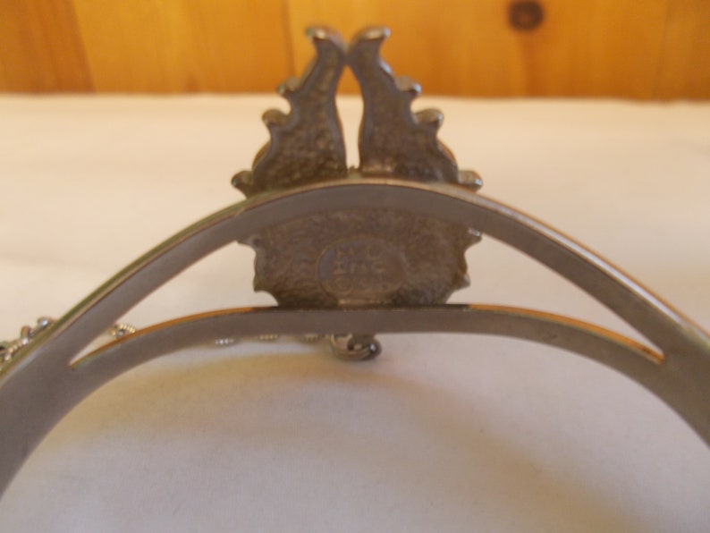 May include: Silver tone bracelet with a decorative design featuring a crown and a chain. The back of the bracelet has a circular stamp with the text "LMC" inside.