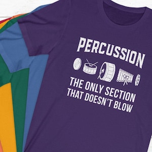 Può includere: Maglietta viola con testo bianco che recita "PERCUSSION THE ONLY SECTION THAT DOESN'T BLOW". La maglietta presenta una grafica di una batteria, piatti e uno xilofono.