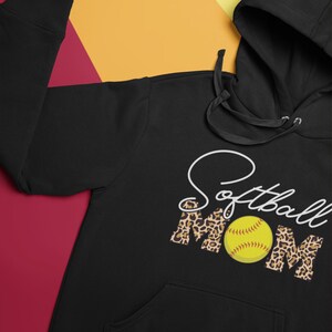 May include: Black hooded sweatshirt with the words "Softball Mom" in white script and leopard print. A yellow softball is in the center of the word "Mom". The sweatshirt has a front pocket and a drawstring hood.