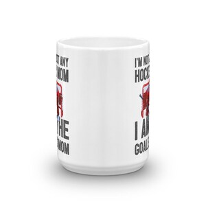 May include: White ceramic coffee mug with a black and white graphic of a hockey goalie in a net. The text on the mug reads "I'm not just a hockey mom, I am the goalie mom" on the left side and "I'm not just a hockey mom, I am the goalie" on the right side.
