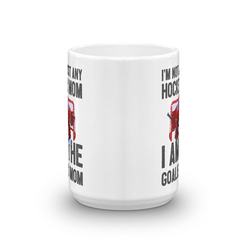 May include: White ceramic coffee mug with a black and white graphic of a hockey goalie in a net. The text on the mug reads "I'm not just a hockey mom, I am the goalie mom" on the left side and "I'm not just a hockey mom, I am the goalie" on the right side.