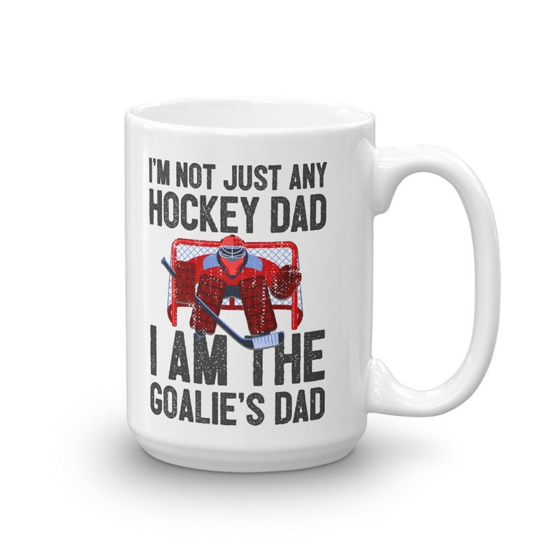 May include: White ceramic mug with a hockey theme. The mug features the text "I'M NOT JUST ANY HOCKEY DAD I AM THE GOALIE'S DAD" in black, with a graphic of a hockey goalie in red and blue gear in front of a net.