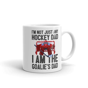 May include: White ceramic mug with a hockey-themed design. The mug features the text "I'M NOT JUST ANY HOCKEY DAD I AM THE GOALIE'S DAD" in black, with a graphic of a hockey goalie in red and blue gear in front of a net.