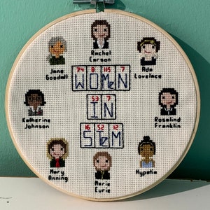 May include: A white embroidery hoop with a cross-stitch design featuring portraits of women in STEM fields. The design includes the text "WOMEN IN STEM" with the atomic numbers for the elements in the word "STEM".