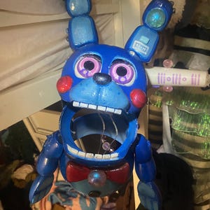 May include: A blue bunny-shaped figurine with a wide open mouth, red cheeks, and a red bow tie. The bunny has large, expressive eyes and two upright ears with lights. The figurine is likely a character from a video game or animated series.