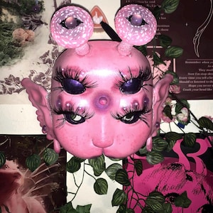 May include: A pink, handmade doll head with four eyes, two large and two small, and two mushroom-shaped antennae. The doll has a pink nose and a small mouth.