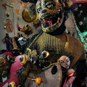 May include: A textured, weathered mask featuring a wide, toothy grin and large, yellow eyes, worn over a dark, mesh-covered garment. The costume is adorned with multiple realistic-looking eyeballs, a prominent orange star, and various glowing red lights.