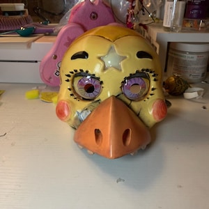 May include: A yellow animatronic character mask with a star on its forehead, black eyelashes, and purple eyes. The mask has a beak-like nose and a toothy grin.