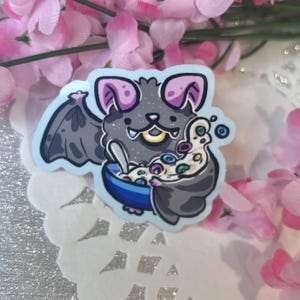 May include: A sticker featuring a cartoon bat with gray fur, purple ears, and a blue bowl. The bat is holding a spoon and a bag filled with colorful, round objects. The background includes pink flowers and a white, decorative surface.