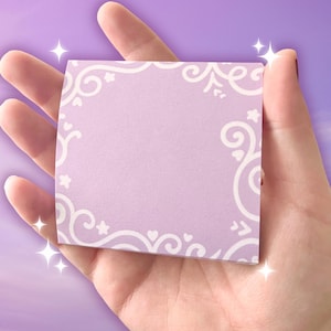 May include: A square, lavender-colored sticky note with a white, ornate border of swirls, hearts, and stars. The note is held in a hand, with a soft focus background. The design is delicate and feminine.