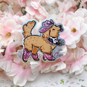 Könnte beinhalten: Ein glitzernder silberner Aufkleber mit einem goldenen Hund, der einen rosafarbenen Cowboyhut und Stiefel trägt. Der Hund trägt ein schwarz-weißes Bandana mit einem Herzmotiv.