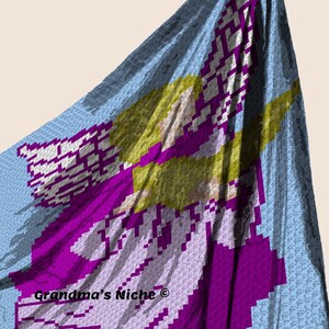 May include: A crochet blanket featuring a purple angel with yellow hair and wings. The blanket is light blue with a white border. The text "Grandma's Niche&reg;" is in the bottom left corner.