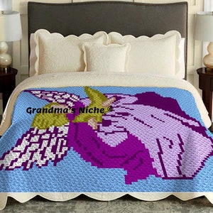 May include: A crochet blanket featuring a purple and white angel with wings, holding a yellow flower. The blanket is on a bed with white pillows and a light blue background.