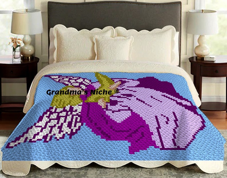 May include: A crochet blanket featuring a purple and white angel with wings, holding a yellow flower. The blanket is on a bed with white pillows and a light blue background.