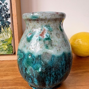 May include: A small ceramic vase with a bulbous shape and a glossy glaze. The upper portion features a speckled light grey and pale green finish, while the lower section transitions into a deep, saturated teal and dark blue with vertical drips.