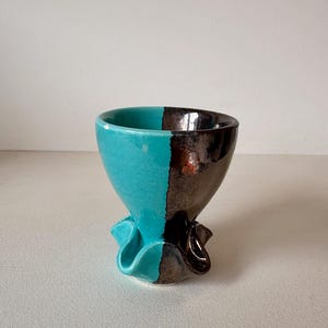May include: A ceramic cup featuring a split design with one side in a glossy turquoise glaze and the other in a dark metallic brown finish. The base of the cup has a distinct ruffled, wavy edge that creates a flared, organic shape at the bottom.
