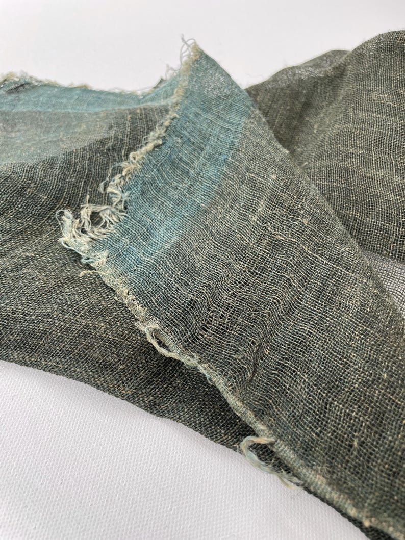 May include: Close-up of a piece of fabric with a frayed edge. The fabric is a dark gray color with a blue-green stripe along the edge. The fabric appears to be loosely woven, with a textured surface.