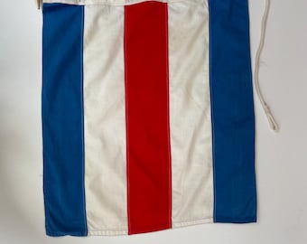 May include: A rectangular nautical flag with a vertical design. The flag features a central red stripe flanked by white stripes, with blue stripes on each side. A wooden dowel and rope are attached to the top edge.