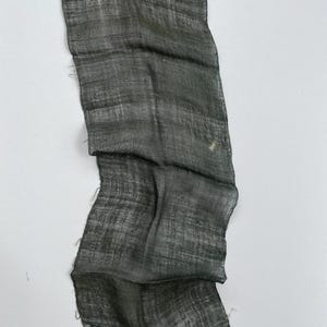 May include: A dark gray, rectangular piece of fabric with a textured weave. The fabric has a slightly wrinkled appearance and frayed edges. The color varies from dark gray to a hint of teal.