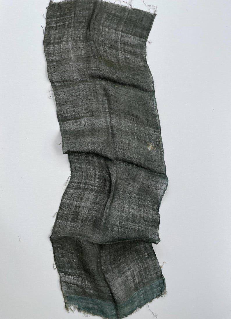 May include: A dark gray, rectangular piece of fabric with a textured weave. The fabric has a slightly wrinkled appearance and frayed edges. The color varies from dark gray to a hint of teal.