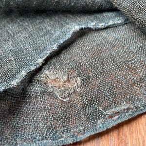 May include: Close-up of a section of teal-colored woven fabric with a visible weave pattern. The fabric has a frayed edge and a small hole, revealing the texture and construction of the material.