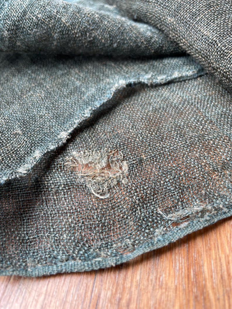May include: Close-up of a section of teal-colored woven fabric with a visible weave pattern. The fabric has a frayed edge and a small hole, revealing the texture and construction of the material.